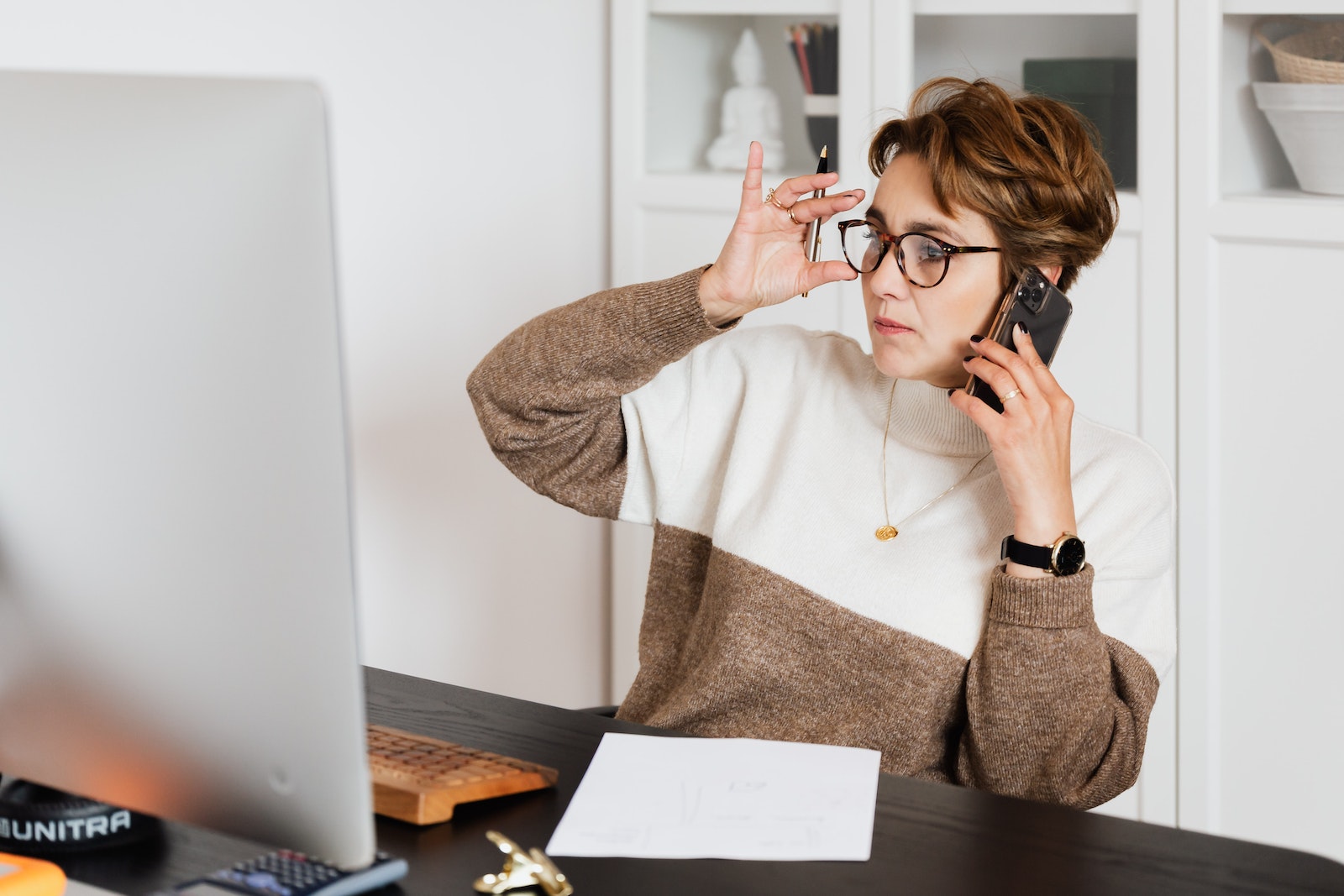 Serious good looking woman in glasses talking on smartphone and looking at computer screen with pen in hand and sheet of paper on desk